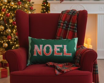 May include: A deep red armchair with a green cushion displaying the word "NOEL" in red. A red and black tartan blanket is draped over the chair. A Christmas tree with gold and red baubles is in the background.