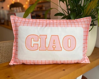 May include: Rectangular pink pillow with a grid pattern and the word "CIAO" in orange block letters. The pillow is on a wooden surface. A plant in a white pot is in the background.