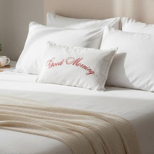 Puede incluir: Una cama con sábanas blancas, almohadas y una manta beige. Una almohada rectangular blanca con las palabras "Good Morning" bordadas en rojo está colocada en la cama, sugiriendo un ambiente acogedor en el dormitorio.
