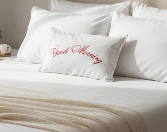May include: A bed dressed with white bedding and a beige throw. A white rectangular pillow with the words "Good Morning" embroidered in red script is placed on the bed. The scene suggests a comfortable and inviting bedroom.