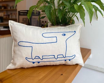 May include: Cream-coloured rectangular cushion featuring a blue embroidered outline of a dog on a skateboard. The dog has a single eye and a long body. The cushion is on a wooden surface with a green plant in the background.