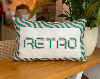 May include: Rectangular cushion featuring the word "RETRO" in green and pink block letters on a white panel. The cushion has a green and pink wavy border. It rests on a wooden surface, with a plant visible in the background.