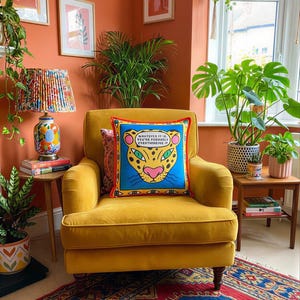 May include: A mustard yellow velvet armchair with a cushion displaying a leopard face and the text "WHATEVER IT IS, YOU'RE PROBABLY OVERTHINKING IT." The room has orange walls, a patterned rug, and potted plants.