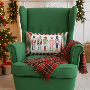 Puede incluir: Un sillón verde con un cojín rectangular con figuras de cascanueces. Una manta a cuadros rojos y verdes está sobre la silla. Los árboles de Navidad y los regalos envueltos están en el fondo, creando una escena festiva.