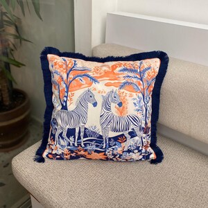 May include: Square cushion with a savanna scene featuring two zebras, in orange, blue and white. It has a navy blue fringe trim. The cushion is placed on a neutral-toned sofa. The design is reminiscent of a safari.
