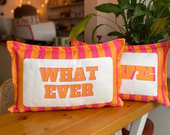 May include: Two rectangular cushions with orange and pink stripes. Each cushion has a white centre with the words "WHAT EVER" in orange block letters. The cushions are on a wooden surface.