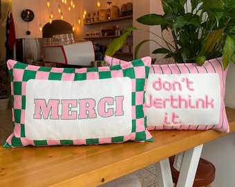 May include: Two rectangular cushions with pink and green chequered borders. One cushion says "MERCI" in pink letters, the other says "don't overthink it." The cushions are on a wooden surface.