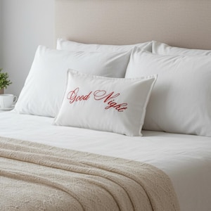 May include: A bed with white pillows and a beige headboard. A white pillow with the words "Good Night" embroidered in red is centered on the bed. A beige knitted throw blanket is draped over the foot of the bed.