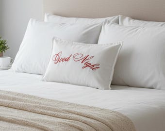 May include: A bed with white pillows and a beige headboard. A white pillow with the words "Good Night" embroidered in red is centered on the bed. A beige knitted throw blanket is draped over the foot of the bed.