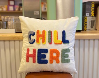 May include: A white cushion with the words "CHILL HERE" embroidered in colourful yarn. The letters are outlined in black and filled with different colours, including red, orange, yellow, green, and blue.