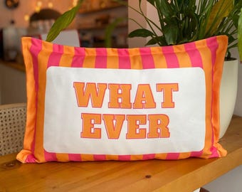 May include: A rectangular cushion with a white central panel featuring the orange text "WHAT EVER". The cushion has a pink and orange striped border. It is displayed on a wooden surface.