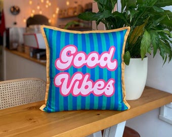 May include: A square cushion with a blue and green striped design, featuring the words "Good Vibes" in pink lettering. The cushion has a yellow border and is placed on a wooden surface.