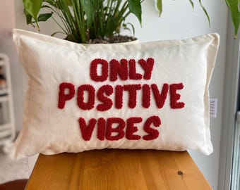 May include: A cream-coloured rectangular cushion embroidered with the words "ONLY POSITIVE VIBES" in red. The cushion is placed on a wooden surface, with a potted plant in the background. The cushion has a tag that says "RAHAN".