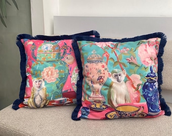 May include: Two decorative cushions with a whimsical design. The cushions feature a monkey, floral patterns, and teacups on a pink and turquoise background. Each cushion has a navy blue fringe border.