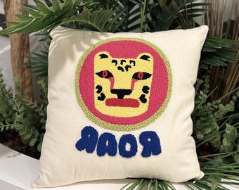 May include: A white cushion with an embroidered tiger face in yellow, pink and green, and the word "ROAR" in blue.