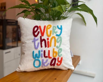 May include: White cushion with the phrase "everything will be okay" embroidered in rainbow colours.