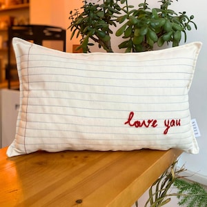 Könnte beinhalten: Ein weißes Kissen mit blauen Linien und den Worten "love you" in rot gestickt. Das Kissen liegt auf einer Holzoberfläche.