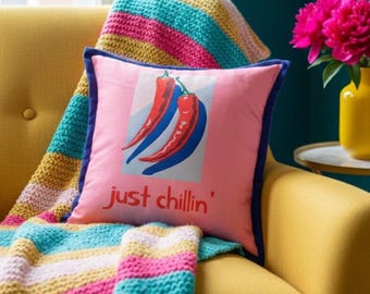 May include: A pink cushion with a blue trim and the words "just chillin'" in red. The cushion features a graphic of two chili peppers in red and blue. A colourful knitted blanket is draped over a yellow sofa.