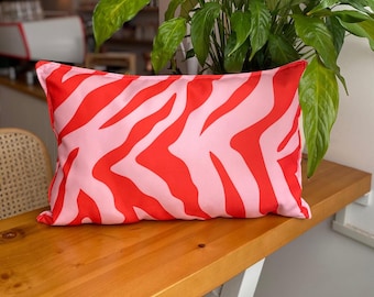 May include: A rectangular cushion featuring a pink base with a striking red zebra stripe design. The cushion is placed on a wooden surface, with a plant in the background. The design adds a vibrant touch to any interior space, with a modern aesthetic.
