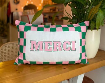May include: A rectangular cushion with a pink and green chequered border. The centre displays the word "MERCI" in pink block letters on a white background. The cushion is placed on a wooden surface.