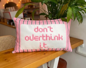 May include: Pink rectangular cushion with green vertical stripes and the words "don't overthink it" in pixelated pink letters. The cushion is on a wooden table, with a plant in the background.