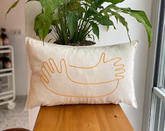 May include: A white cushion with a yellow embroidered design of two hands reaching towards each other.