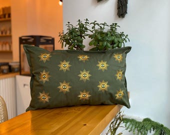 May include: A green cushion with a repeating pattern of gold stars with an eye in the centre. The cushion is on a wooden surface.