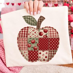 May include: A white t-shirt featuring a large apple-shaped applique made of various fabric patches. The patches include red and white gingham, floral patterns, polka dots, and heart prints. A green gingham leaf and brown stem are attached.