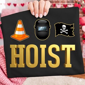May include: Black t-shirt with gold 3D text that reads "HOIST". The shirt features three graphics: an orange and white traffic cone, a black welding mask, and a pirate flag with a skull and crossbones.