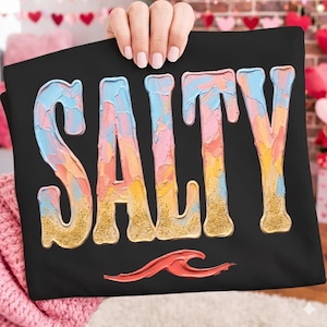 Op de afbeelding: Zwart t-shirt met het woord "SALTY" in grote, kleurrijke, getextureerde letters. De letters zijn geschilderd in tinten blauw, roze, geel en oranje met gouden glitteraccenten. Een rood golfontwerp staat onderaan.