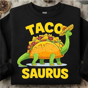 Puede incluir: Sudadera negra con una ilustración de dibujos animados de un dinosaurio verde con forma de taco, con un sombrero. El texto "TACO" está encima del dinosaurio y "SAURUS" debajo, ambos en amarillo.