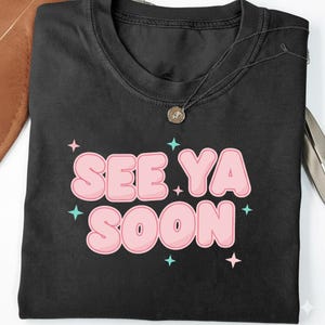 May include: Black t-shirt with the phrase "SEE YA SOON" in bubble letters. The text is pink with a white outline, and there are teal and pink star accents around the text. A silver necklace with a pendant is on the shirt.