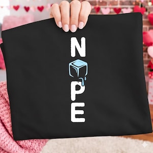 Op de afbeelding: Zwart T-shirt met het woord "NOPE" in witte hoofdletters. De "O" is vervangen door een blauwe ijsblokjesafbeelding met druppelend water. Het shirt wordt door een persoon omhooggehouden.