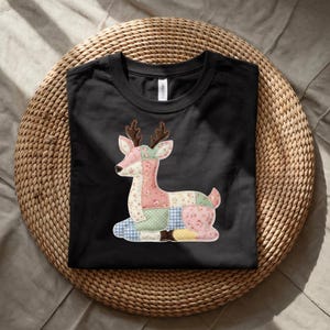 May include: Black t-shirt featuring a patchwork deer design. The deer is composed of various fabric patterns, including floral and gingham prints, in shades of pink, green, blue, and yellow. The shirt is displayed on a woven, circular surface.