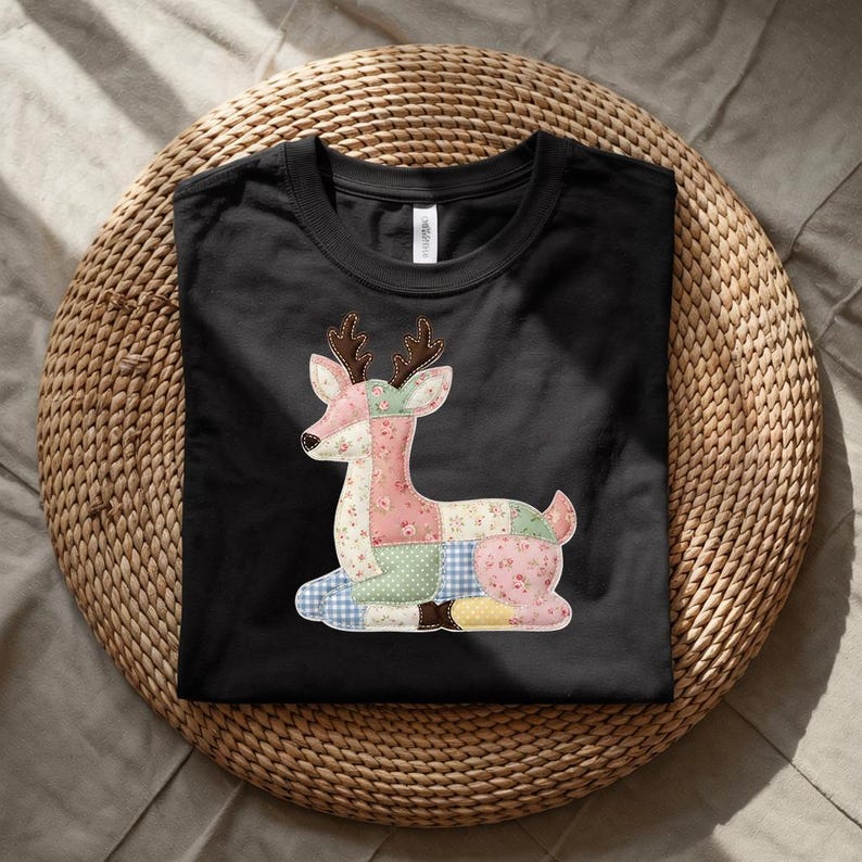 May include: Black t-shirt featuring a patchwork deer design. The deer is composed of various fabric patterns, including floral and gingham prints, in shades of pink, green, blue, and yellow. The shirt is displayed on a woven, circular surface.