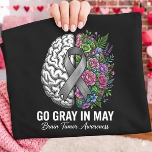 Könnte beinhalten: Schwarzes T-Shirt mit einem Design zur Sensibilisierung für Hirntumore. Das Design zeigt ein Gehirn, ein graues Band und ein Blumenarrangement. Der Text "GO GRAY IN MAY Brain Tumor Awareness" ist darunter gedruckt.