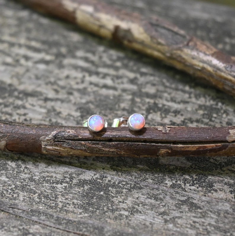 May include: A pair of opal stud earrings. Each earring features a round, iridescent opal stone set in a silver-colored metal. The opals display a mix of pink, orange, and white hues. The earrings are displayed on a wooden branch.