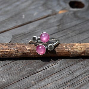 May include: A bypass ring featuring two round, cabochon-cut pink stones and two smaller, smooth silver-toned metal discs. The band is crafted from a silver-toned metal with a textured finish, showcasing a modern design with alternating stone and metal elements.