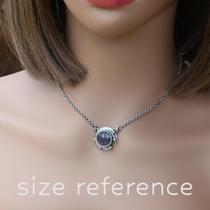 May include: A silver chain necklace with a round pendant featuring a purple gemstone set in a silver bezel. The pendant is surrounded by a decorative silver frame.