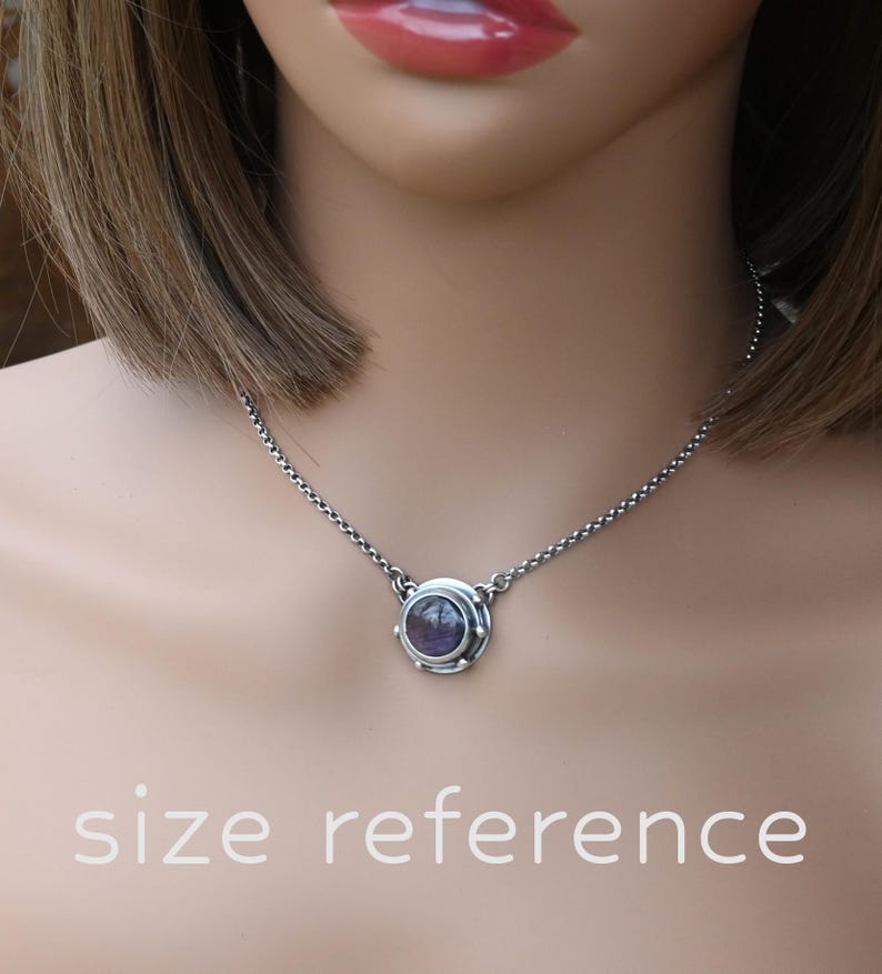 May include: A silver chain necklace with a round pendant featuring a purple gemstone set in a silver bezel. The pendant is surrounded by a decorative silver frame.