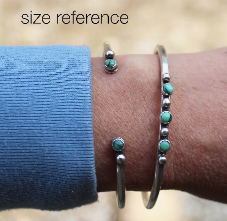 May include: Two silver bracelets with turquoise stones on a person's wrist. The bracelet on the bottom has five turquoise stones and the bracelet on the top has one turquoise stone. The bracelets are on a person's wrist with a blue sleeve.