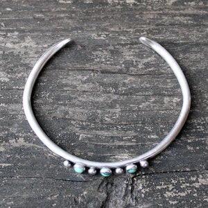 May include: A silver bangle bracelet with turquoise stones set in the metal. The bracelet is open and has a simple design.