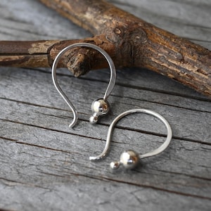May include: A pair of silver-colored, curved earrings. Each earring features a rounded hook design with a small, spherical bead and a larger, polished sphere at the base. The earrings are displayed on a weathered wooden surface.