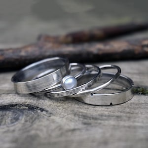 Può includere: Una collezione di anelli d'argento, alcuni con fasce strutturate e uno con una piccola perla bianca rotonda. Gli anelli sono disposti su una superficie di legno invecchiato, creando un'esposizione rustica ed elegante.