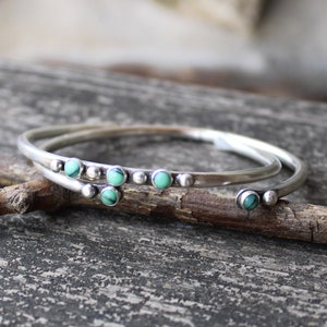 May include: Two silver bangles with turquoise stones. The bangles are stacked on top of each other and are lying on a piece of wood.