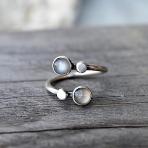 May include: A silver-toned ring with a wrap-around design. The ring features three light-colored, round, and oval-shaped stones. The ring is set against a weathered, gray wooden surface. The ring is a piece of jewelry.