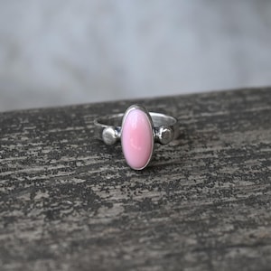 May include: A silver ring with a pink oval gemstone set in the centre. The ring has two small silver balls on either side of the gemstone.