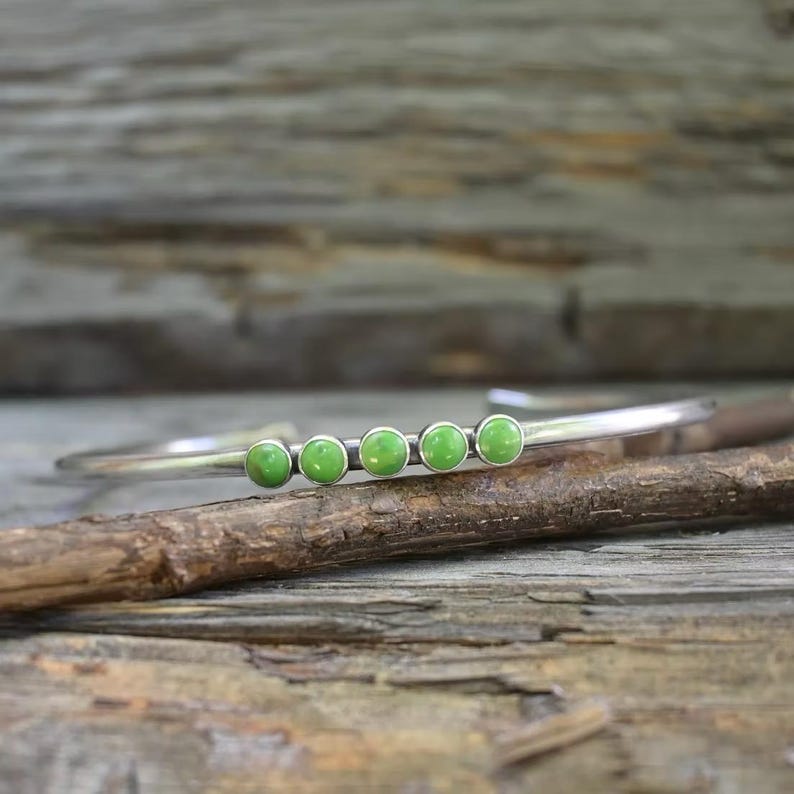 May include: A silver-toned cuff bracelet featuring five small, round, light green cabochon stones set in a row along the center of the band. The metal has a smooth, polished finish, and the stones are evenly spaced, creating a simple, linear design.