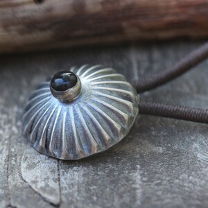 May include: A silver pendant with a black gemstone set in the center. The pendant has a textured, fluted design and is attached to a brown cord.