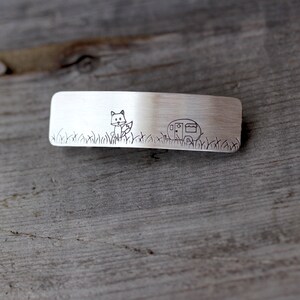 May include: A silver metal hair clip with a fox and a camper engraved on it. The fox is facing the camper and there is grass engraved around them.